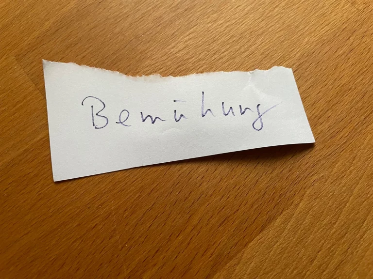 Handgeschriebener Notizzettel mit dem Wort „Bemühung“ auf einem Holztisch.