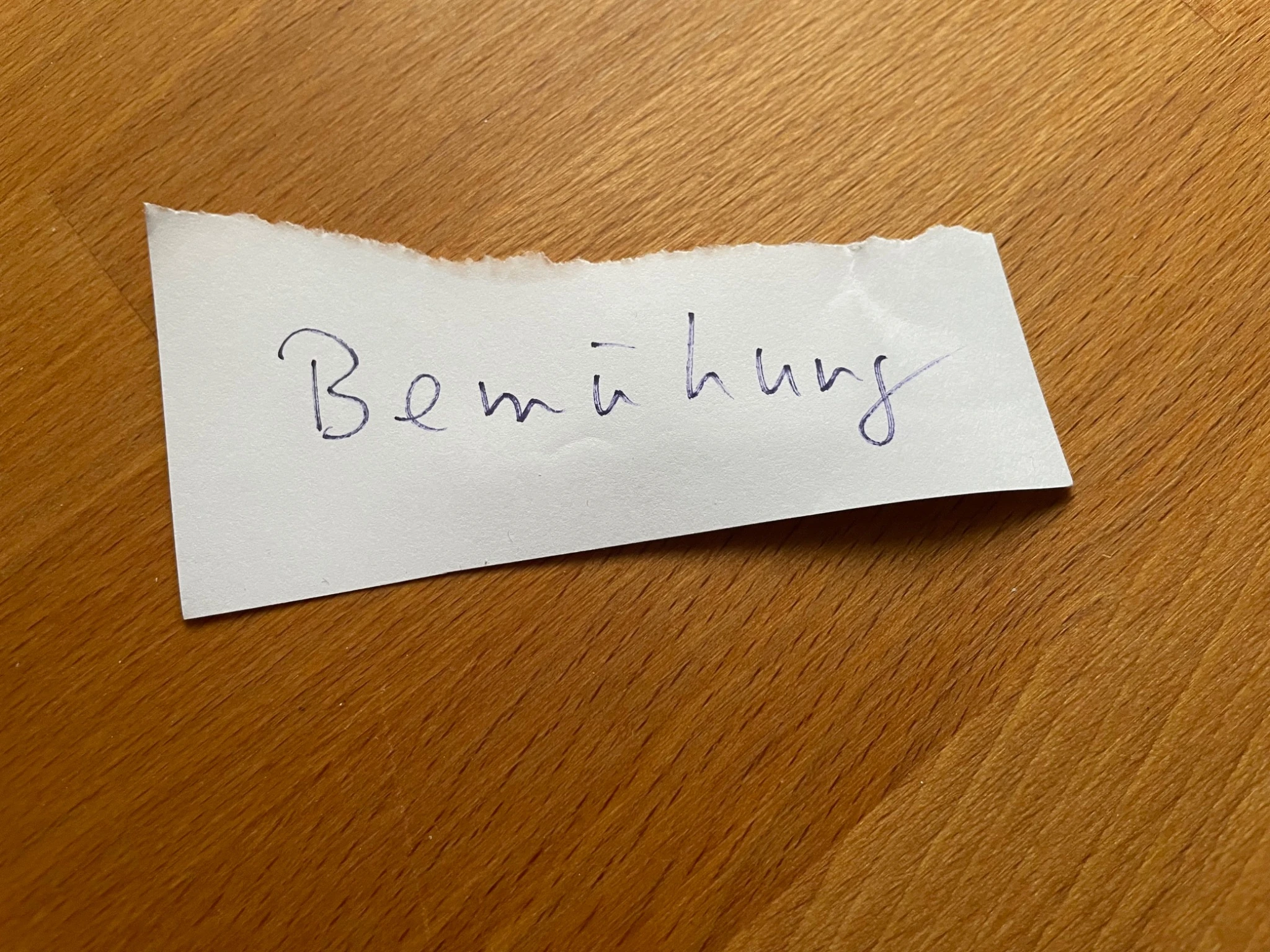 Handgeschriebener Notizzettel mit dem Wort „Bemühung“ auf einem Holztisch.
